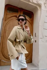 Trench corto de color beige, con cuello de solapa, manga larga con hombreras y trabillas con botones. Cuenta con cierre frontal cruzado con botones oscuros y cinturón ajustable del mismo tejido.