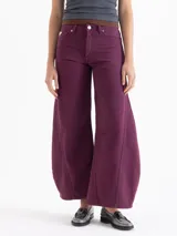 Pantalón de jean con silueta wide leg y diseño redondeado. Presenta pinzas en la zona de las rodillas que acentúan su forma envolvente, cierre frontal con botón metálico, bolsillos laterales y remache decorativo distintivo.