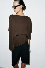 Top tipo capa blanco de punto fluido, con mezcla de viscosa, cuello bote, mangas cortas y bajo asimétrico.