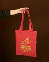 Tote bag de tela color rojo con estampado gráfico en color verde lima que representa una fogata y una persona sentada tomando una bebida caliente, acompañado por el texto 'Invierno Uruguayo'.