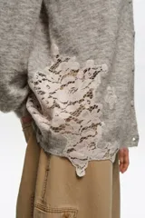 Cardigan gris melange de punto con escote en V y cierre frontal de botones. Presenta aplicaciones de encaje en tono rosado en el hombro izquierdo y en el lateral inferior derecho.