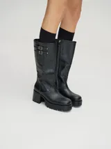 Bota bucanera de cuero vacuno wax color negro, con punta cuadrada, suela de goma con plataforma dentada y taco de madera. Presenta detalle de doble hebilla metálica en la parte superior de la caña.