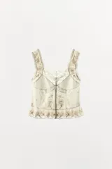 Top de algodón color crema con escote redondo, tirantes anchos y cierre frontal con botones. Presenta detalles de plisados, encaje combinado, bordados florales y aplicaciones de abalorios, con el bajo acabado en ondas.