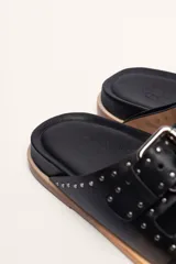Sandalias de cuero color negro con dos tiras anchas sobre el empeine, cada una con hebillas metálicas ajustables y detalles de tachas plateadas. La suela es de cuero con un borde de color madera.
