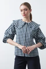 Camisa de algodón con estampado a cuadros vichy en azul y blanco. Presenta un cuello grande con volados, mangas largas con volumen y puños elásticos, y cierre frontal con botones.