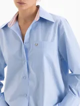 Camisa de corte holgado confeccionada en poplín de algodón. Presenta cuello clásico con interior en tela a rayas a contratono, cierre frontal con botones al tono, bolsillo plaque en el pecho con detalle de remache metálico en forma de corazón y mangas largas con puños estructurados.