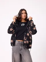 Campera reversible de diseño holgado, con un lado en tejido sherpa estampado con motivos florales en tonos negro y beige, y el otro lado en nylon negro. Cuenta con cierre frontal y cordones ajustables en el ruedo.