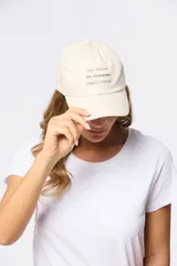 Gorro con visera de lona color beige con bordado frontal de texto en tonos rosado, gris y celeste.