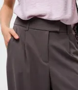 Pantalón de sastrería modelo barrel, de tiro alto y corte amplio con piernas anchas. Presenta pliegues frontales, trabillas para cinturón y recortes decorativos en la parte delantera.