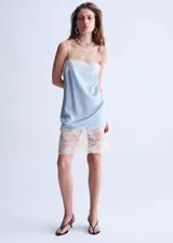 Vestido corto de tejido satinado color celeste, con diseño recto, escote recto y tirantes finos. Presenta detalles de encaje blanco en el escote y en el ruedo.