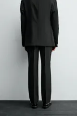 Pantalón de vestir negro con pliegues frontales, corte regular fit, bolsillos laterales y traseros de doble vivo con botón. Cierre frontal cruzado con cremallera y gancho metálico.