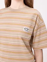 Remera de corte holgado con diseño de rayas horizontales en tonos beige, rosa y verde oliva. Presenta cuello redondo, mangas cortas y un bolsillo frontal pequeño con el logo de la marca.