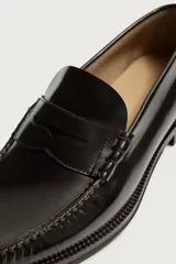 Mocasines de vestir tipo loafer para hombre, confeccionados en cuero marrón oscuro con acabado brillante. Presentan diseño clásico de antifaz en el empeine, costuras visibles en relieve y suela plana con un ligero tacón.