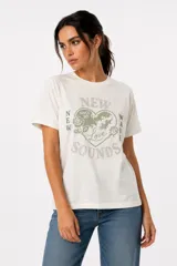 Remera de algodón color blanco off-white, de manga corta, cuello redondo a la base y corte holgado. Presenta un estampado frontal con diseño de corazón en tonos marrones y azules, con detalles de strass y texto.