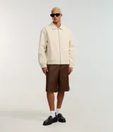 Campera boxy de poliéster color beige, con cuello inglés, cierre frontal de cremallera, mangas largas con elástico en los puños, bolsillos laterales y detalles de recortes.