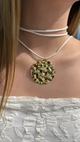 Gargantilla tipo choker compuesta por un cordón blanco de doble vuelta y un dije circular grande de metal dorado con textura irregular y efecto espejo.