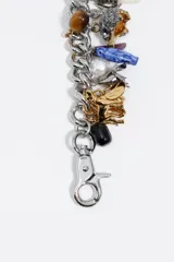 Pulsera de cadena metálica con múltiples dijes colgantes de diversos materiales, incluyendo figuras de animales, piedras naturales, elementos de resina y piezas metálicas en tonos dorados y plateados. Cuenta con cierre de mosquetón.
