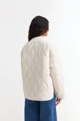 Campera acolchada de diseño liviano en color beige, con cuello redondo, cierre frontal con cremallera y dos bolsillos delanteros tipo parche. Presenta un patrón de costuras onduladas en toda la prenda.