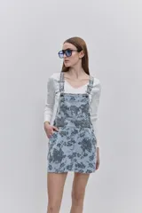 Jumper de jean corto, de color celeste claro con estampado floral en tonos oscuros. Presenta tirantes regulables con hebillas metálicas, bolsillo pectoral y bolsillos laterales funcionales.