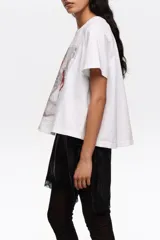 Remera blanca de algodón, de corte línea A con vuelo, escote redondo y mangas cortas. Presenta un estampado frontal con diseño de zapatillas de ballet en tonos grises y lazos rojos.
