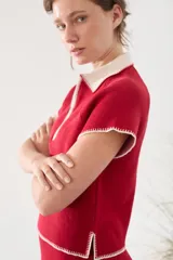 Top tejido de punto color rojo con cuello polo blanco, medio cierre frontal y detalles de costuras visibles en contraste en los bordes de las mangas y el ruedo.