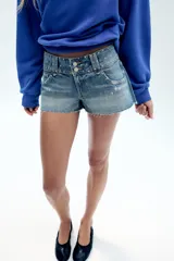 Shorts de denim de tiro bajo con cinco bolsillos, detalle de tachas, aplicación de brillos, bajo deshilachado y cierre frontal con cremallera y botones metálicos.