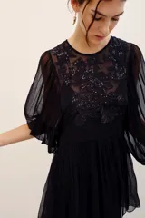 Vestido corto negro con transparencias, mangas largas amplias y detalles de bordados florales con lentejuelas en el frente. Cuenta con un lazo ajustable en la cintura y falda fluida.