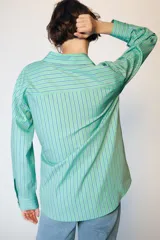 Camisa de manga larga con cuello solapa, cierre frontal con botones y diseño cruzado con lazada lateral en el mismo tejido. Presenta un estampado de rayas verticales en tonos verdes y azules.