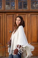 Poncho tejido de color blanco, con flecos en los bordes y detalles de bordado en crochet a lo largo de la abertura frontal. Cuenta con un cierre de cordones con borlas en el cuello.