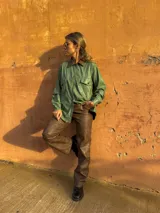 Camisa verde de cuero sintético con cuello marrón, cierre frontal con botones, dos bolsillos con solapa en el pecho y cinturón marrón.