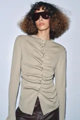 Chaqueta de punto color beige claro, manga larga y cuello redondo. Presenta un detalle de frunces verticales ajustados por botones a tono en el centro frontal.