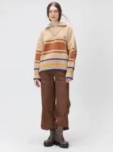 Pullover de punto con textura aterciopelada, diseño oversize, cuello polo con cierre frontal parcial y detalles de pespuntes en contraste. Presenta rayas horizontales en tonos tierra y un parche bordado con la letra M en el pecho.