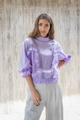 Blusa lila de cuello alto y manga 3/4 con elástico en el bajo. Presenta volados verticales en el frente.