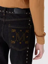 Jeans de corte ancho (wide leg) y tiro medio, confeccionados en denim de algodón negro con costuras contrastantes en color ocre. Presenta múltiples botones metálicos frontales y tachas metálicas aplicadas a lo largo de los laterales y la pretina.