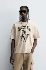 Camiseta de corte relaxed fit, color beige claro con efecto lavado, cuello redondo y manga corta. Estampado frontal en negro con texto "SUNSET ERA" y un caballo galopando.