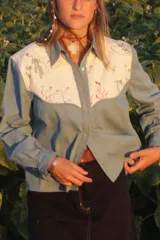 Camisa de denim estilo western con canesú delantero en contraste de color crema con estampado de cactus y motivos desérticos. Presenta cuello clásico, cierre frontal con botones y mangas largas.