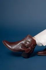 Bota corta de cuero color chocolate, con taco cuadrado y hebilla metálica decorativa en el lateral. Presenta un diseño clásico con punta redondeada.