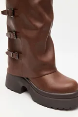 Bota alta marrón de diseño slouchy con plataforma track y múltiples hebillas metálicas decorativas en la parte lateral exterior de la caña.