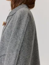 Capa de tejido de punto en color gris melange, con diseño abierto y cierre frontal mediante un botón tipo duffle de madera con detalle en cuero. Presenta bolsillos laterales amplios y bordes con costuras a la vista.