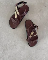 Sandalias planas de cuero color marrón con tiras cruzadas decoradas con apliques metálicos dorados. Cuentan con ajuste de pulsera al tobillo mediante hebilla metálica.