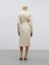 Vestido camisero midi color crema, de lino y viscosa, con cuello chal asimétrico, manga larga con puños abotonados, cierre frontal con botones y cinturón con hebilla forrada.