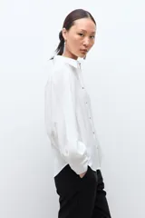 Camisa blanca de manga larga con puños abotonados y fruncidos, hombros fruncidos y cierre frontal con botones.