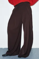 Pantalón de tiro medio con pierna ancha, confeccionado en tela de color marrón oscuro. Presenta pliegues frontales decorativos, bolsillos laterales ocultos en la costura y cierre frontal con cremallera, botón y ganchos metálicos.