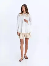 Camisa blanca vaporosa con cuello mao, botonadura frontal oculta y mangas largas abullonadas con puños elásticos. Presenta un diseño de paneles fruncidos y detalles bordados con motivos geométricos dorados en la parte superior delantera.