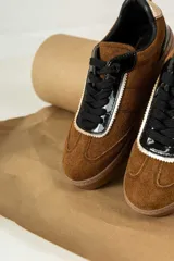 Championes urbanos de gamuza color marrón, con detalles en charol negro y bordes dorados. Presentan cordones negros y suela de goma al tono.