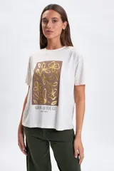 Remera de manga corta color crudo con estampa frontal rectangular en tonos marrones y dorados con diseño floral y la frase 'Grow as you go'.