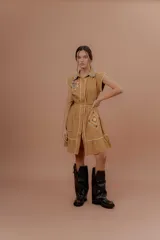 Vestido corto beige de algodón con cuello camisero y cierre frontal de botones. Presenta mangas cortas y detalles bordados a contratono en la zona del pecho y en los bolsillos, además de un volado en el ruedo.