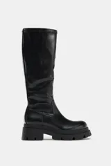 Bota de caña alta con suela de plataforma dentada y punta redonda. Presenta un diseño minimalista con costuras visibles y cierre de cremallera lateral.
