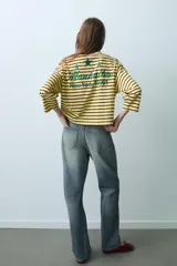 Camiseta oversize de manga larga con diseño de rayas horizontales amarillas y negras. Presenta cuello redondo y un detalle de texto bordado en el centro del pecho.