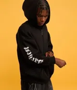 Saco tipo hoodie con cierre frontal de cremallera, color negro, corte boxy y capucha. Presenta estampa de letras en blanco en ambas mangas y un colgante de cruz metálico en el tirador de la cremallera principal.